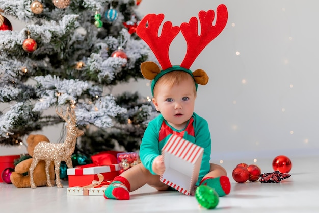 Baby in einem grünen Body mit roten Hirschhörnern auf dem Kopf spielt mit Geschenkboxen in der Nähe des Weihnachtsbaums. Kind in einem Weihnachtszwergkostüm. Winter-Neujahrskonzept. Urlaubsware. Platz für Text