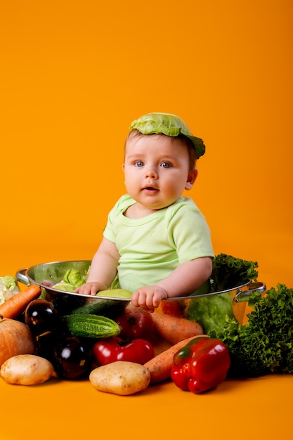 Baby in einem grünen Bodysuit sitzt in einer Metallschale mit Gemüse. landwirtschaftliches Produktkonzept