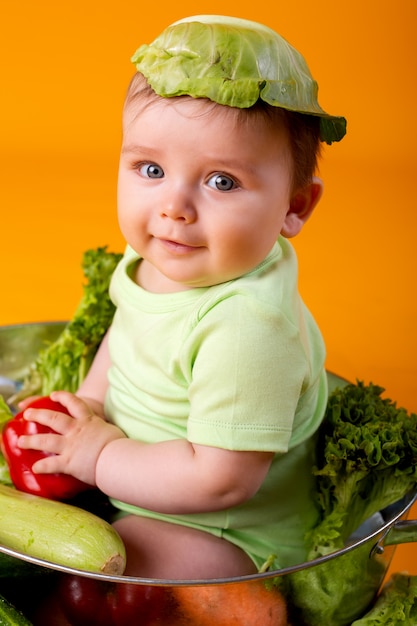 Baby in einem grünen Bodysuit sitzt in einer Metallschale mit Gemüse. landwirtschaftliches Produktkonzept