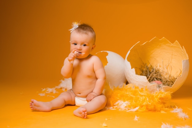 Baby in einem Hühnerkostüm mit Federn und Ostereiern auf einer orange Wand, Platz für Text. das Konzept von Ostern