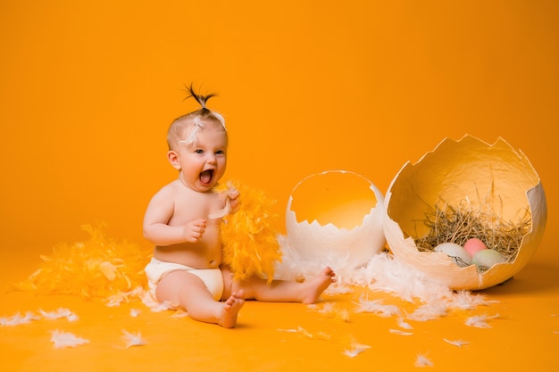 Baby in einem Hühnerkostüm mit Federn und Ostereiern auf einer orange Wand