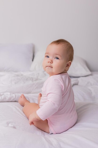 baby in einem rosa bodysuit sitzt mit dem rücken zur kamera in einem bettchen auf schneeweißer bettwäsche