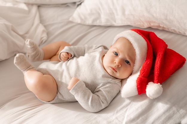 Baby in einem weißen Body und einer Weihnachtsmütze liegt im Bett neben dem Weihnachtsbaum. charmantes blauäugiges Kind. Winterurlaub, lustige Momente, Kinderweihnachten. Platz für Text. Foto in hoher Qualität