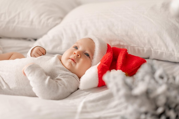 Baby in einem weißen Body und einer Weihnachtsmütze liegt im Bett neben dem Weihnachtsbaum. charmantes blauäugiges Kind. Winterurlaub, lustige Momente, Kinderweihnachten. Platz für Text. Foto in hoher Qualität