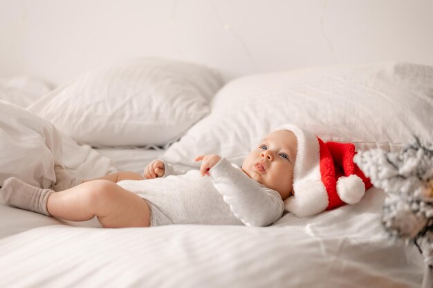 Baby in einem weißen Body und einer Weihnachtsmütze liegt im Bett neben dem Weihnachtsbaum