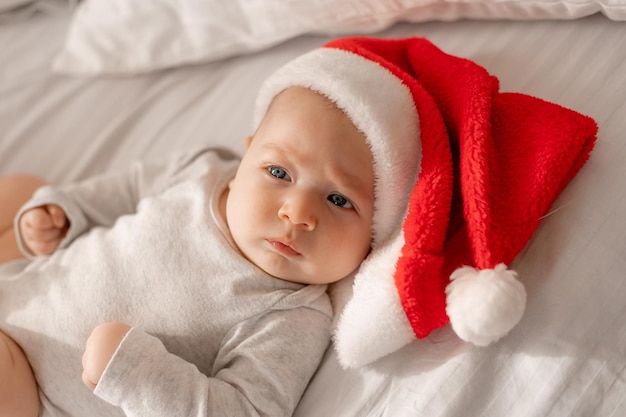 Baby in einem weißen Body und einer Weihnachtsmütze liegt im Bett neben dem Weihnachtsbaum