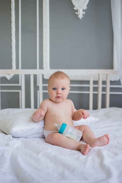 Baby in einer Windel, die auf dem Bett mit einer Flasche Formel sitzt