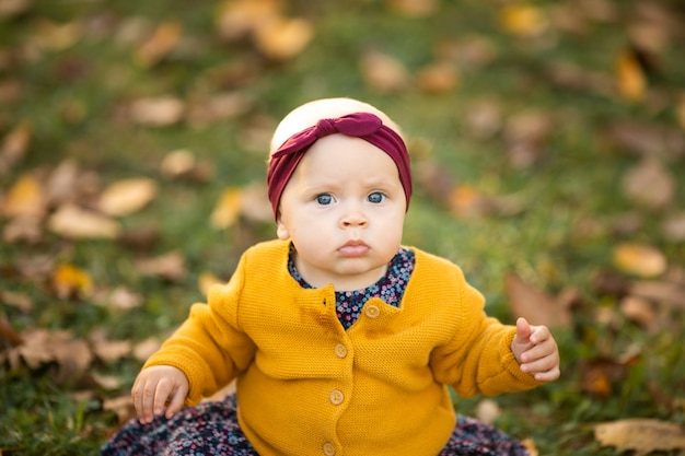 Baby in gelber Jacke und rotem Stirnband, die auf dem Gras sitzen und im Herbstlaub spielen.