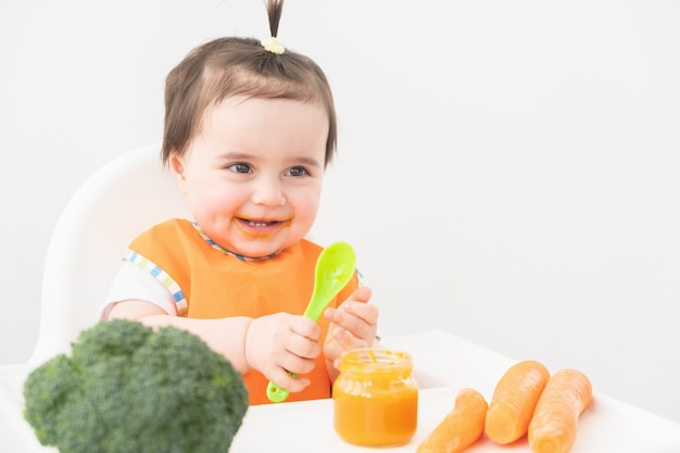 Baby in orangeem Lätzchen, das in einem Kinderstuhl sitzt, der Gemüsepüree auf weißem Hintergrund isst.