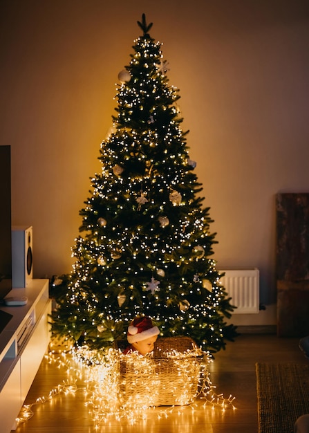 Baby in Weihnachtsmütze, sitzt unter einem Weihnachtsbaum mit Lichtern zu Hause.