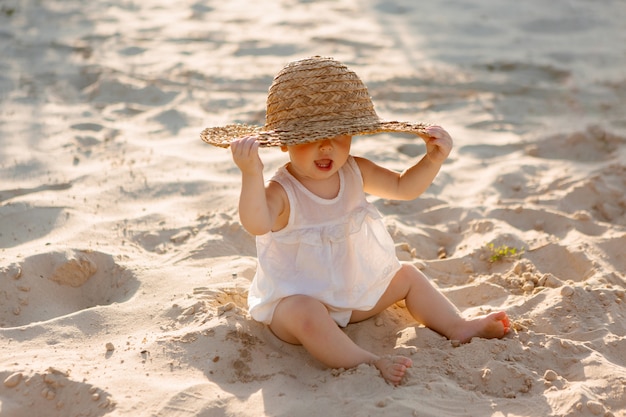 Baby in weißen Kleidern und einem Strohhut sitzt im Sommer auf dem weißen Sand am Strand