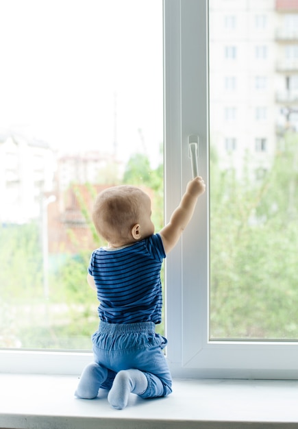 Baby Junge sitzt auf der Fensterbank und versucht, das Fenster zu öffnen, indem es am Griff zieht