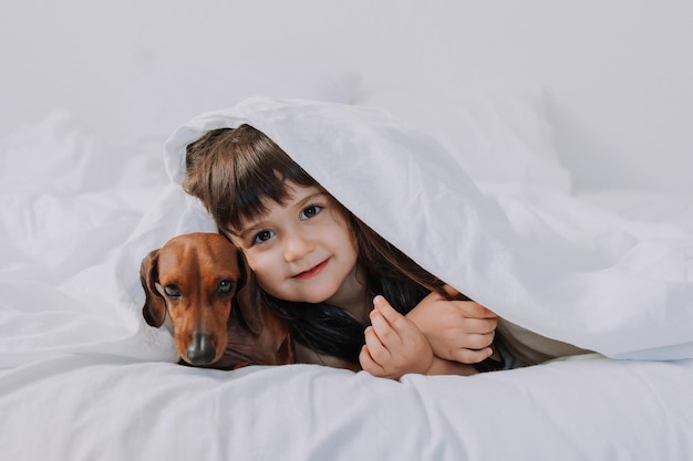 Baby kleines Mädchen umarmt Dackelhund zu Hause im Bett. weiße Bettwäsche. Liebe für Haustiere