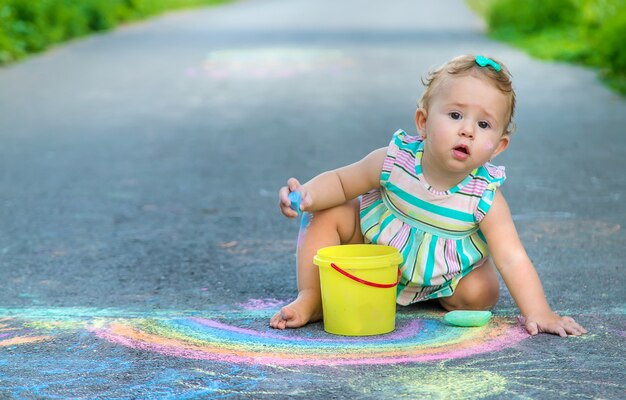 Baby malt mit Kreide einen Regenbogen auf den Bürgersteig. Selektiver Fokus. Natur.