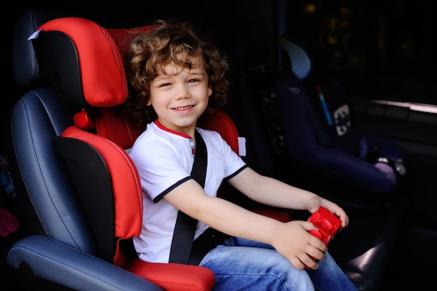 Baby mit dem gelockten Haar, das in einem Kinderautositz mit Spielzeugauto in den Händen sitzt