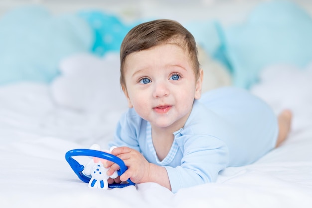 Baby mit einem Nagetier zum Zahnen oder einer Rassel auf dem Bett zum Schlafen, gesundes, glückliches kleines Baby in einem blauen Body spielen