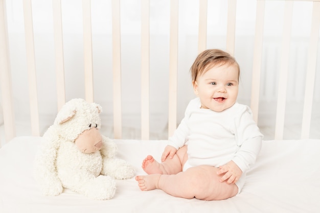 Baby sechs Monate sitzt in einer Krippe in einem weißen Body mit Spielzeug Teddybären