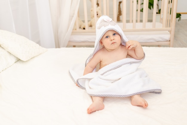 Baby sitzt auf einem weißen Bett in einem weißen Handtuch nach dem Baden im Badezimmer zu Hause