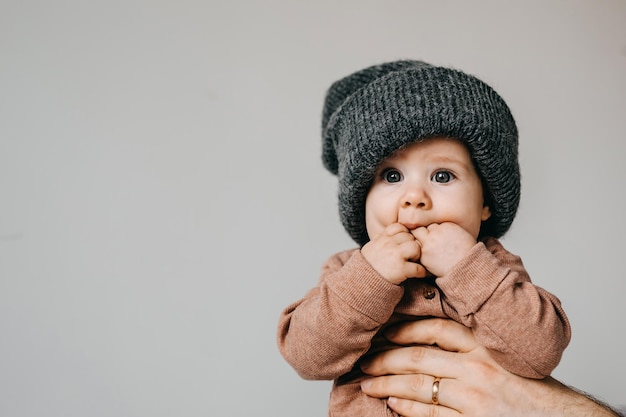 Baby trägt eine graue übergroße Strickmütze und lutscht Finger auf weißem Wandhintergrund