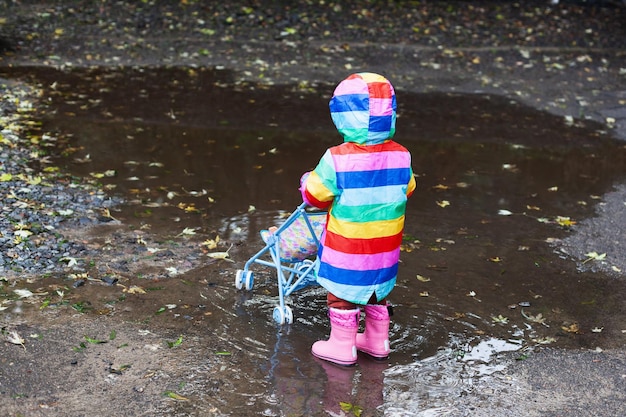 Baby trägt rosa Gummistiefel und bunten Regenbogenmantel und spielt in einer Herbstpfütze
