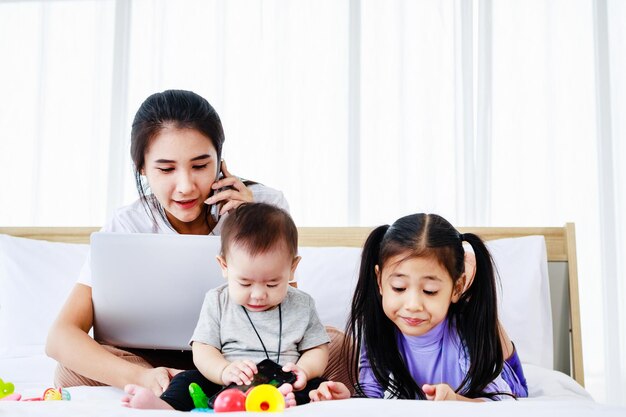 Baby und kleine Tochter spielen Spielzeug auf dem Bett, während die beschäftigte freiberufliche Mutter am Laptop arbeitet