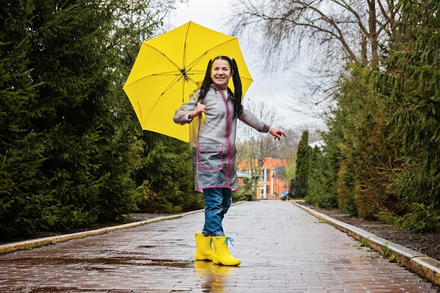 Babyboomer Fröhliche Seniorin im gelben Regenmantel mit gelbem Regenschirm, die springt und das Leben im Park genießt