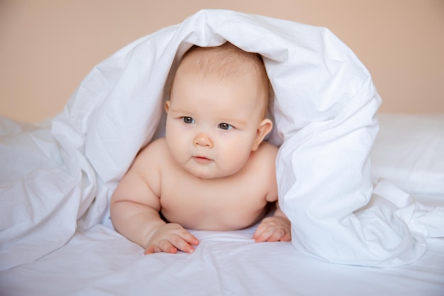 Babyjunge in einer Windel liegt auf einem weißen Laken, bedeckt mit einer Decke im Schlafzimmer auf dem Bett