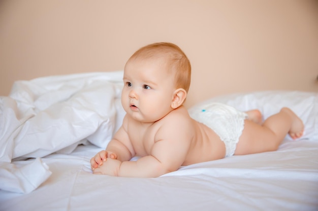 Babyjunge in einer Windel liegt auf einem weißen Laken, bedeckt mit einer Decke im Schlafzimmer auf dem Bett