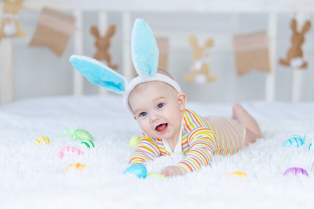 Babyjunge mit Hasenohren auf seinem Kopf liegend auf dem Bett mit Ostereiern, niedliches lustiges lächelndes kleines Baby.