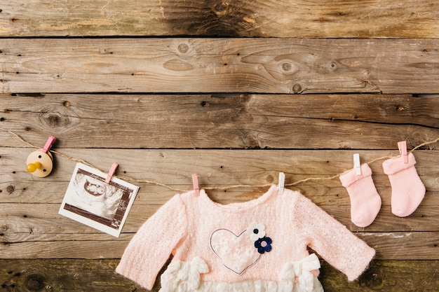 Foto babykleidung; socken; schnuller- und sonographiebild, das an der wäscheleine mit wäscheklammern gegen hölzerne wand hängt