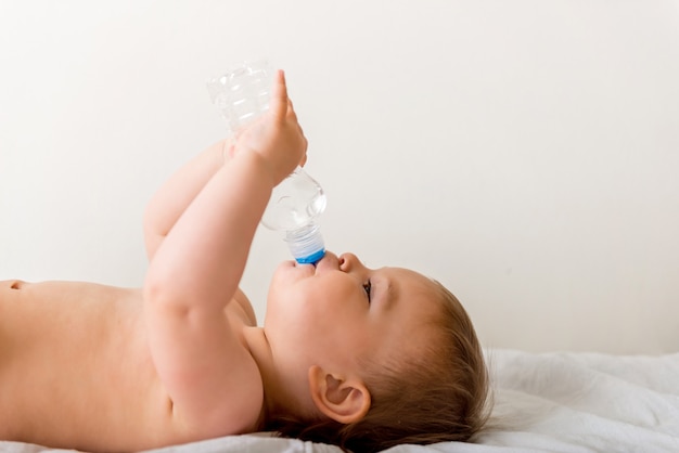 Babykleinkind sitzt auf dem weißen Bett, lächelt und trinkt Wasser von der Plastikflasche