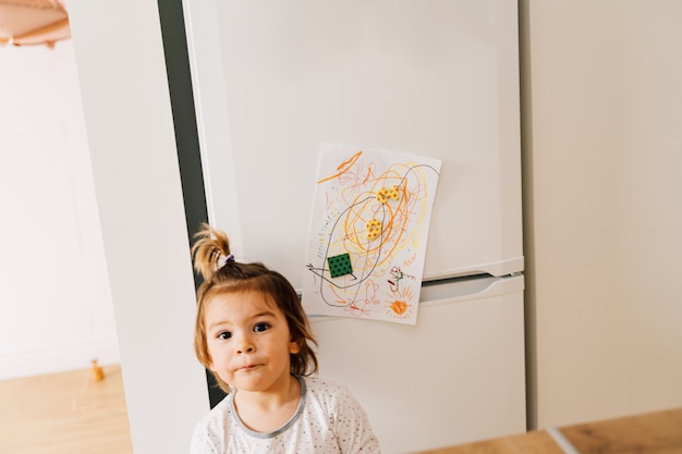 Babykleinkindmädchen, das ihr Kunstwerk am Kühlschrank mit Magneten aufhängt