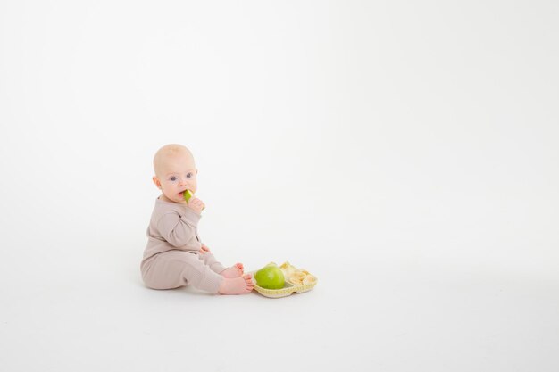 Babymädchen, das ein Stück Apfel isst, sitzend auf einem Studioaufnahmen mit weißem Hintergrund