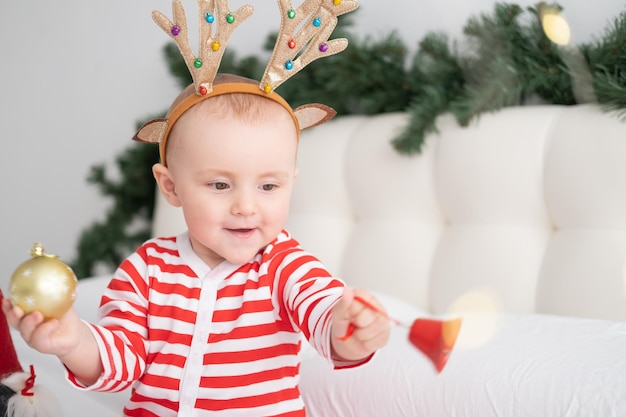 Babymädchen im gestreiften Body mit Hirschhörnern auf dem Kopf, die mit Weihnachtsglocke auf dekoriertem Bett spielen