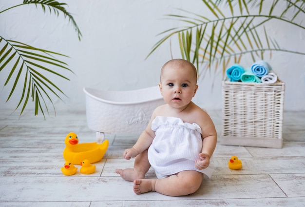 Babymädchen in einem Badetuch sitzt in der Nähe einer weißen Badewanne auf dem Boden und beobachtet die Kamera