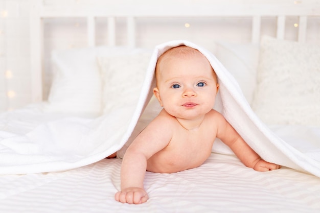 Babymädchen in einem Handtuch nach dem Baden in einer Krippe auf einem weißen Baumwollbett, das auf dem Bauch liegt und in der Neugeborenenhygiene des Kindergartens lächelt