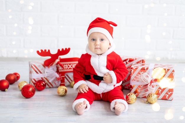 Babymädchen in einem roten Weihnachtsmannkostüm sitzt mit Kisten mit Geschenken und Weihnachtsspielzeug auf weißem Hintergrund