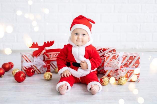 Babymädchen in einem roten Weihnachtsmannkostüm sitzt mit Kisten mit Geschenken und Weihnachtsspielzeug auf weißem Hintergrund