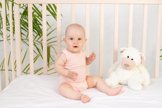 Babymädchen in einer Krippe in einem rosa Body und einem Teddybären sechs Monate