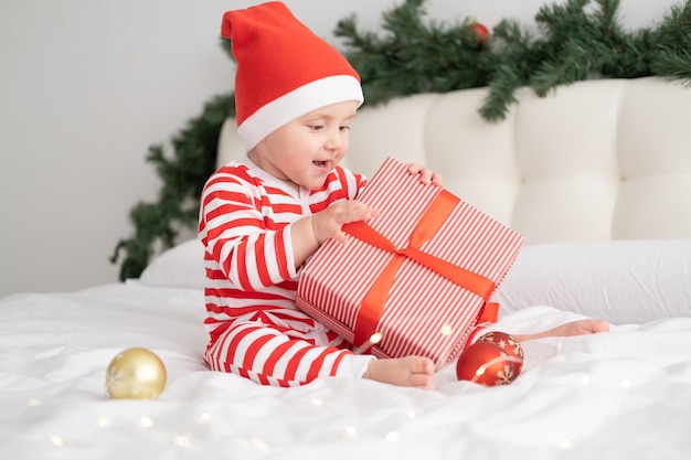 Babymädchen in gestreiftem Body und Weihnachtsmütze, das mit Geschenkbox auf weihnachtlich dekoriertem Schlafzimmer spielt