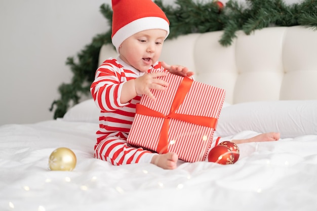 Babymädchen in gestreiftem Body und Weihnachtsmütze, das mit Geschenkbox auf weihnachtlich dekoriertem Schlafzimmer spielt