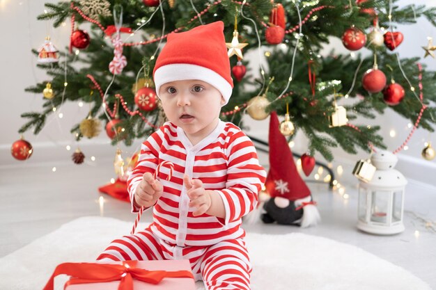 Babymädchen in gestreiftem Body und Weihnachtsmütze spielt mit Geschenkbox in der Nähe des Weihnachtsbaums