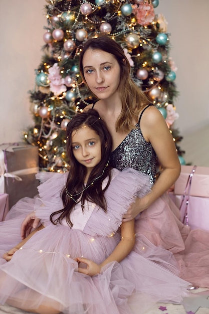 Babymädchen mit großen Augen und einer Mama in einem rosa Kleid sitzt am Weihnachtsbaum