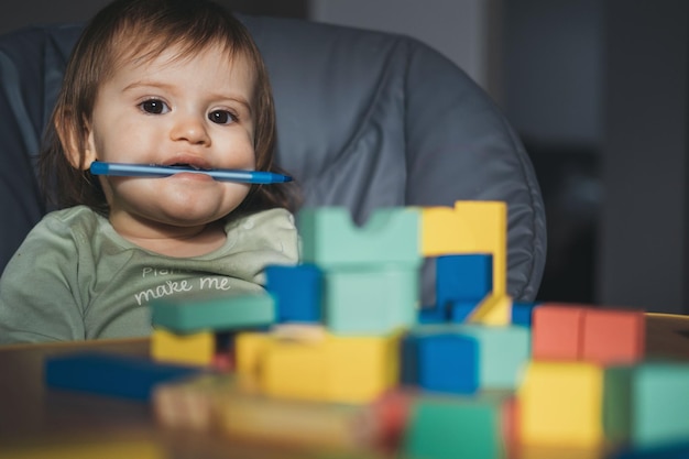 Babymädchen sitzt auf dem Hochstuhl und hält einen Stift im Mund, der auf das Kamerakind schaut