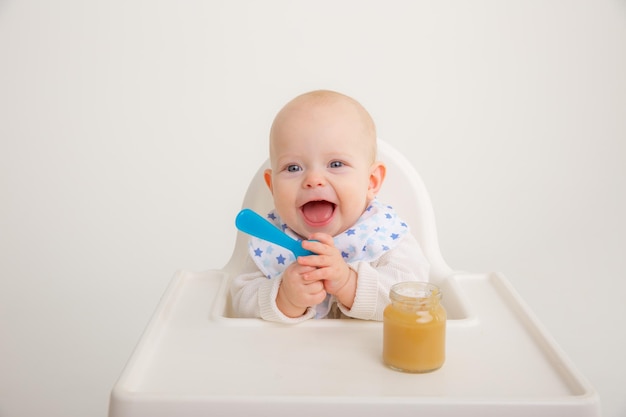 Babymädchen sitzt auf einem Hochstuhl, hält Löffel und isst Fruchtpüree