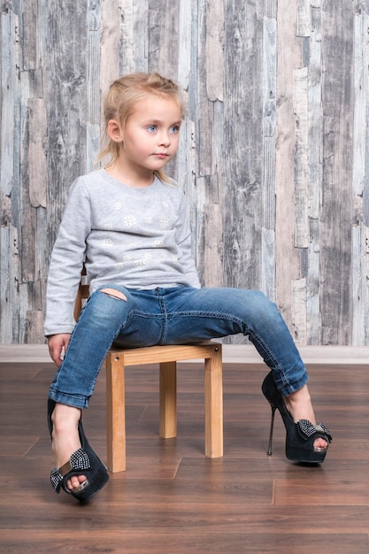 Babymädchen sitzt auf einem Holzstuhl, die Beine tragen große Schuhe ihrer Mutter