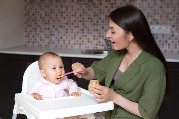 Babynahrung Ein Baby in einem Hochstuhl isst Gemüsepüree aus einem Löffel Mama füttert das Baby aus einem Glas Küche Lifestyle