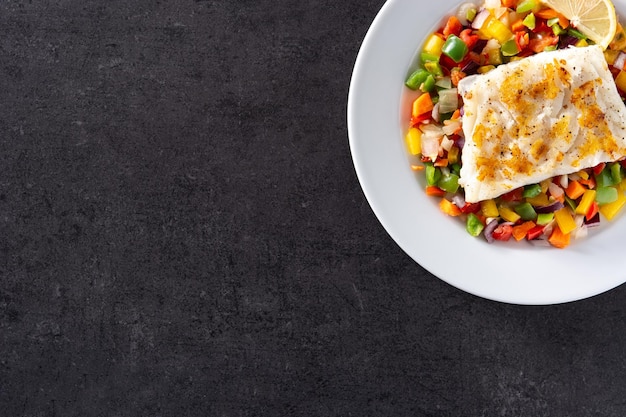 Bacalhau grelhado com legumes no prato sobre fundo preto de pedra