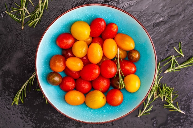 Bacia de tomates de cereja coloridos (vermelho, granada e amarelo), frescos e crus. Com gotas de água e alecrim.