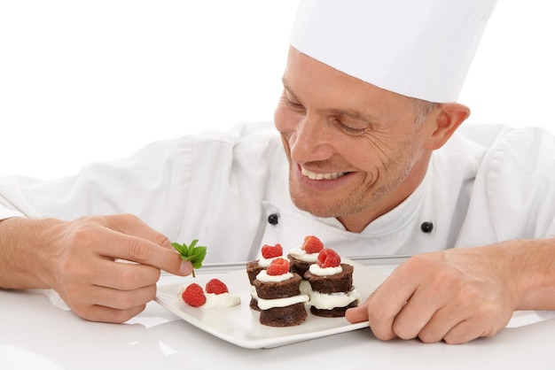 Foto backen, lächeln und koch mit einem dessert für die gastronomie isoliert auf weißem hintergrund kochprofi und mann, der einen schokoladenkuchen und obst auf einem teller für einen food-service vor dem hintergrund überzieht